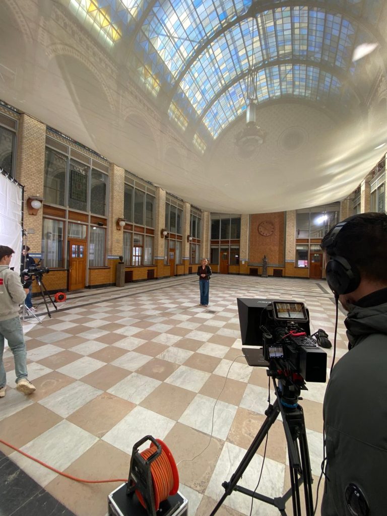 Camera crew filming a historical scene in a glass-roofed hall for Ieder uur een woord, een daad.